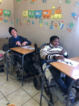 Two students using supportive wheelchairs in a colorful classroom at the Lighthouse Stimulation Centre